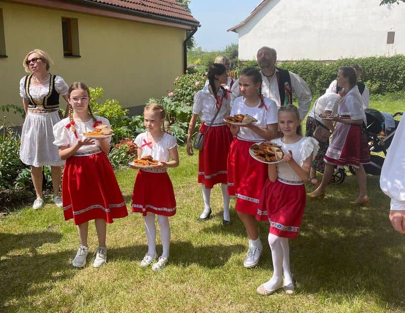 Děti v krojích drží talíře s jídlem, v pozadí dospělí v krojích