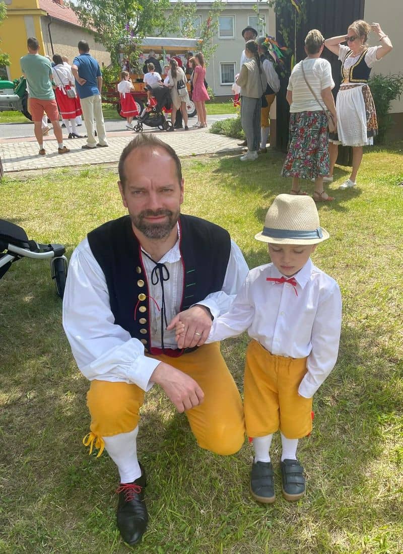 Muž a chlapec v krojích na trávě, v pozadí další lidé
