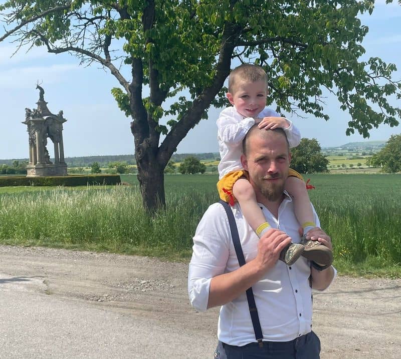 Muž nese dítě na ramenou, v pozadí strom a socha