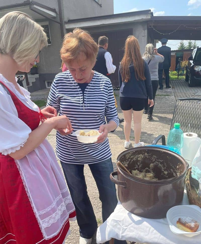 Žena v kroji podává jídlo z hrnce jiné ženě