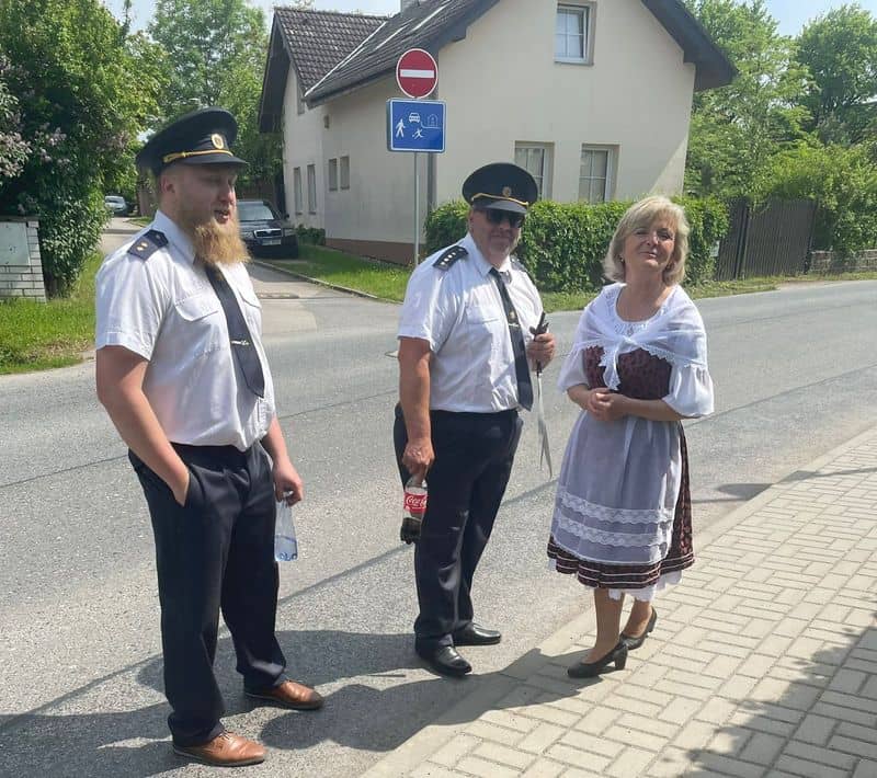 Dva muži v uniformách a žena v kroji stojí na chodníku