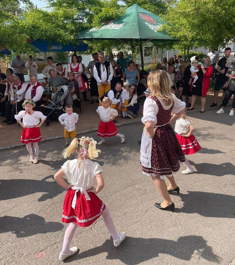 Děti a dospělí v krojích tančí na venkovní akci, přihlíží skupina lidí