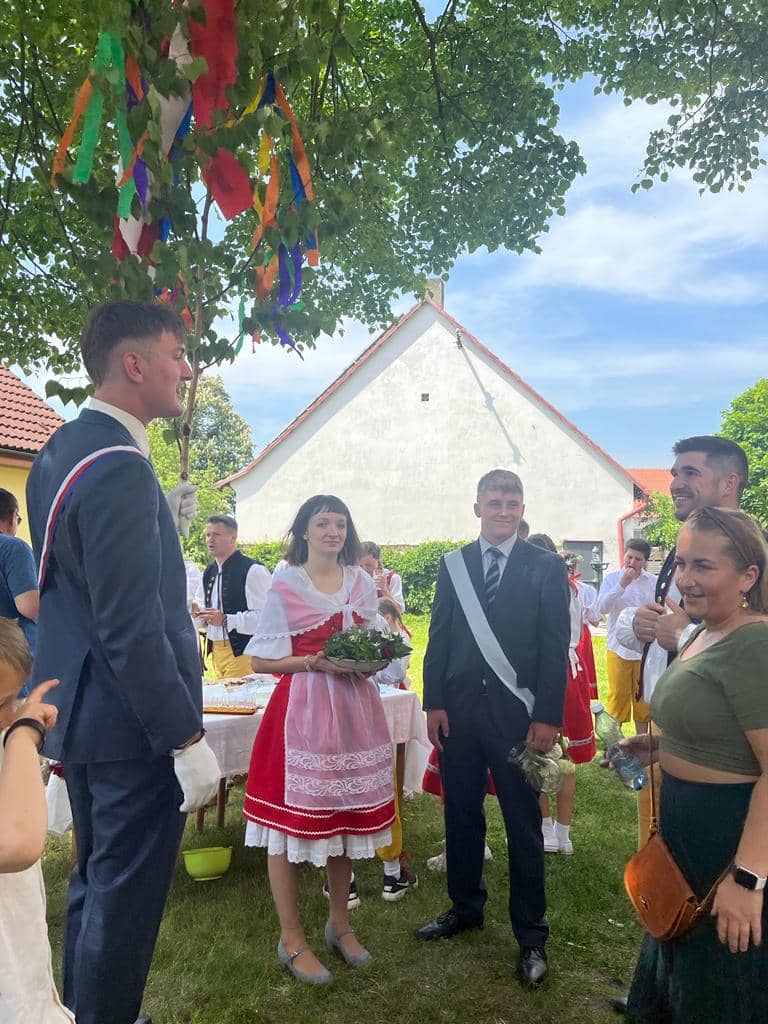 Skupina lidí v krojích a oblecích pod stromem s barevnými stuhami