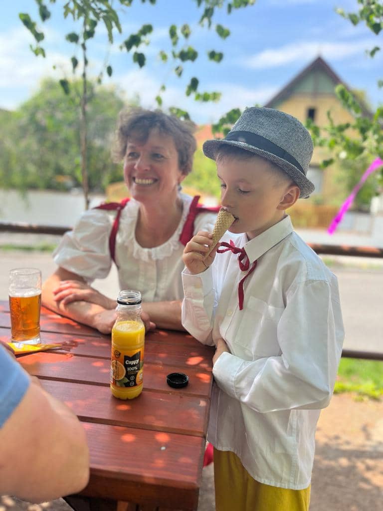 Žena v kroji a chlapec v klobouku jí zmrzlinu u stolu venku