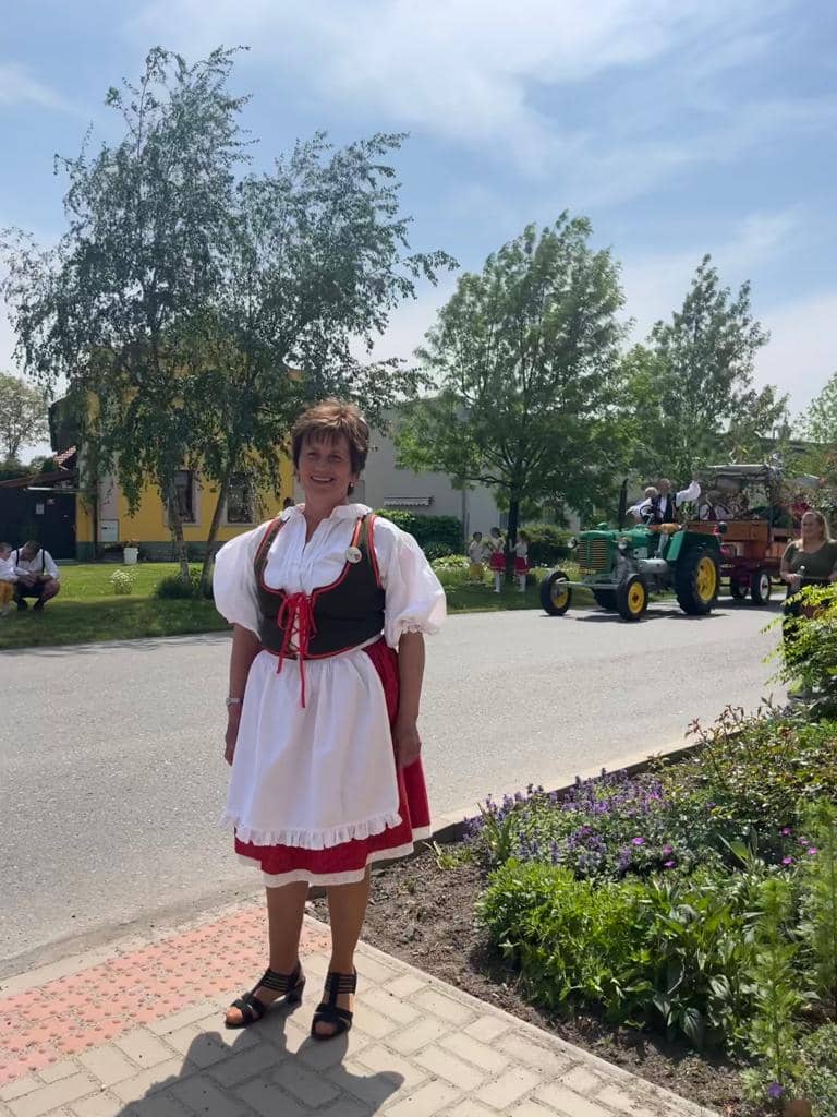 Žena v kroji stojí u silnice, v pozadí traktor s vlečkou
